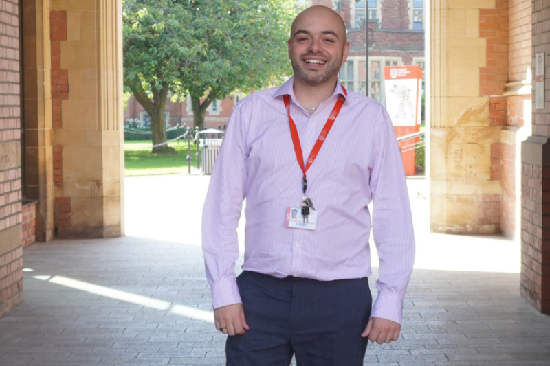 Bald smiling male standing in an arched walkway in front of a sunlit green area. He is wearing a light pink open buttoned shirt and dark grey trousers, as well as a red lanyard around his neck.